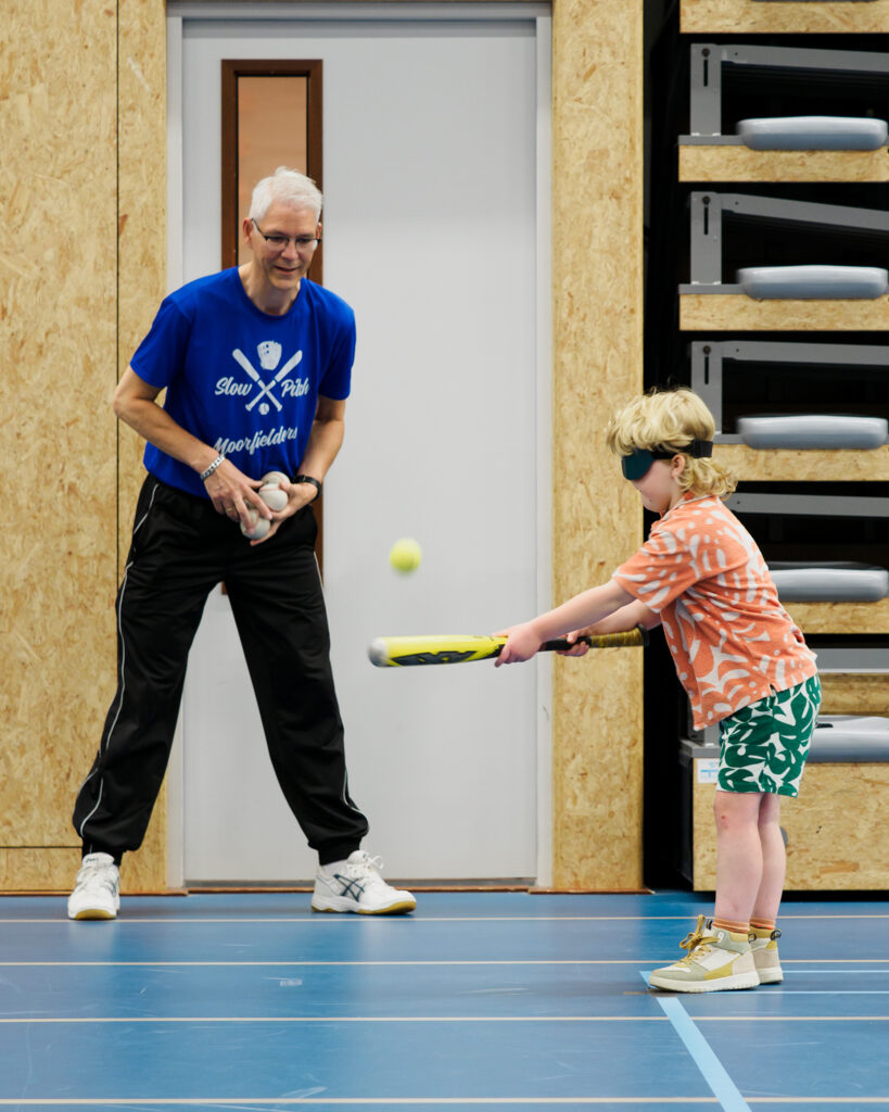 Op de afbeelding een jonge deelnemer van de multisportdag die met een knuppel de bal slaat. Daarachter staat de baseballtrainer instructies te geven.