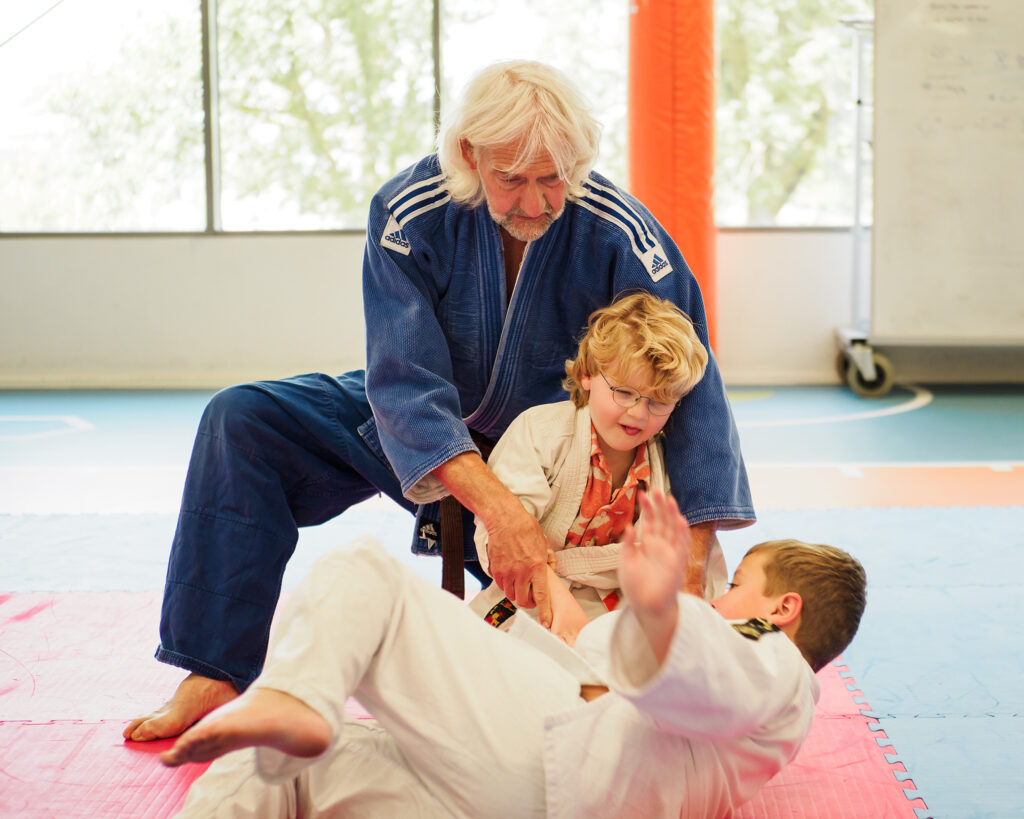 Op de afbeelding de judo instructeur die twee jonge deelnemers begeleidt in een judo gooi.