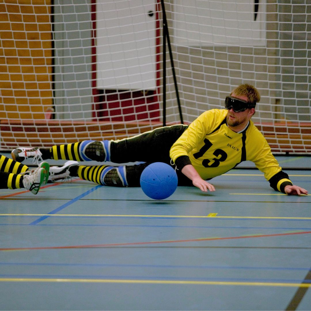 goalball speler met geel shirt duikt voor een blauwe bal.
