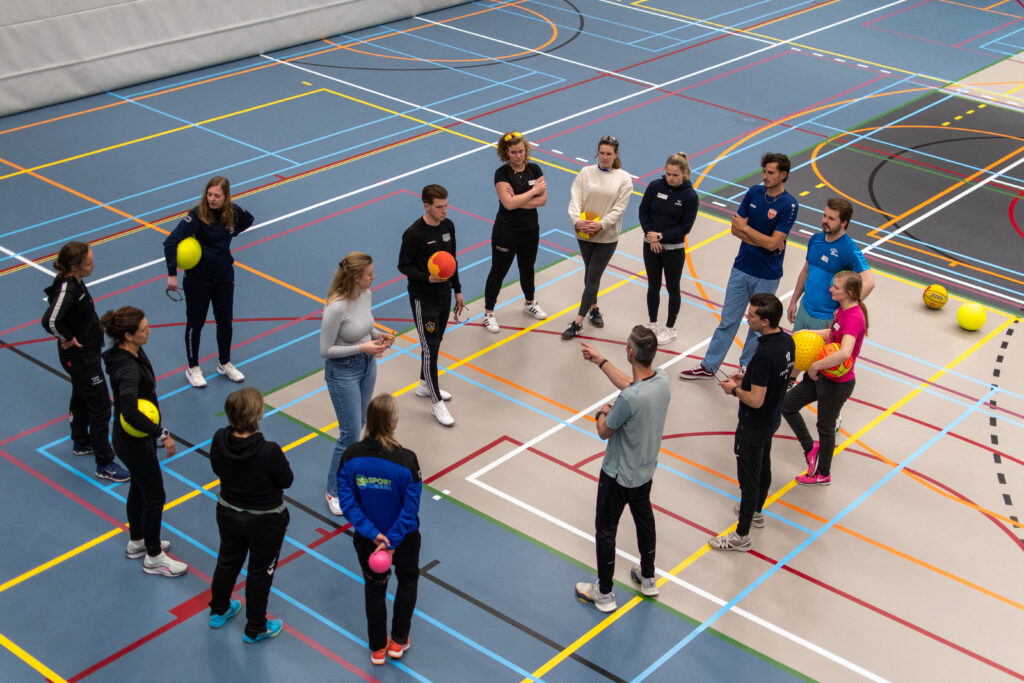 de deelnemers van de trainingsdag van boven gefotografeerd terwijl ze uitleg krijgen in de sportzaal
