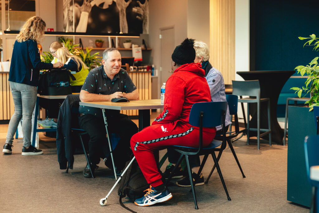 Meerdere mensen aan een tafel in een koffietent. 1 van de mensen heeft een taststok.