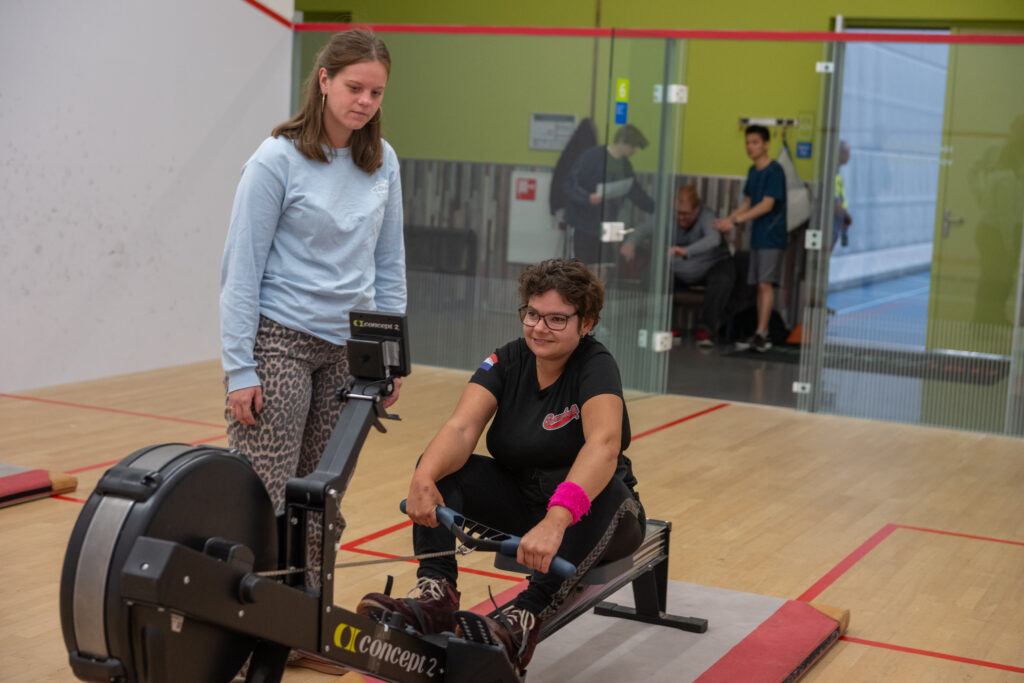 Deelneemster multisportdag op een ergometer met naast haar een instructeur.