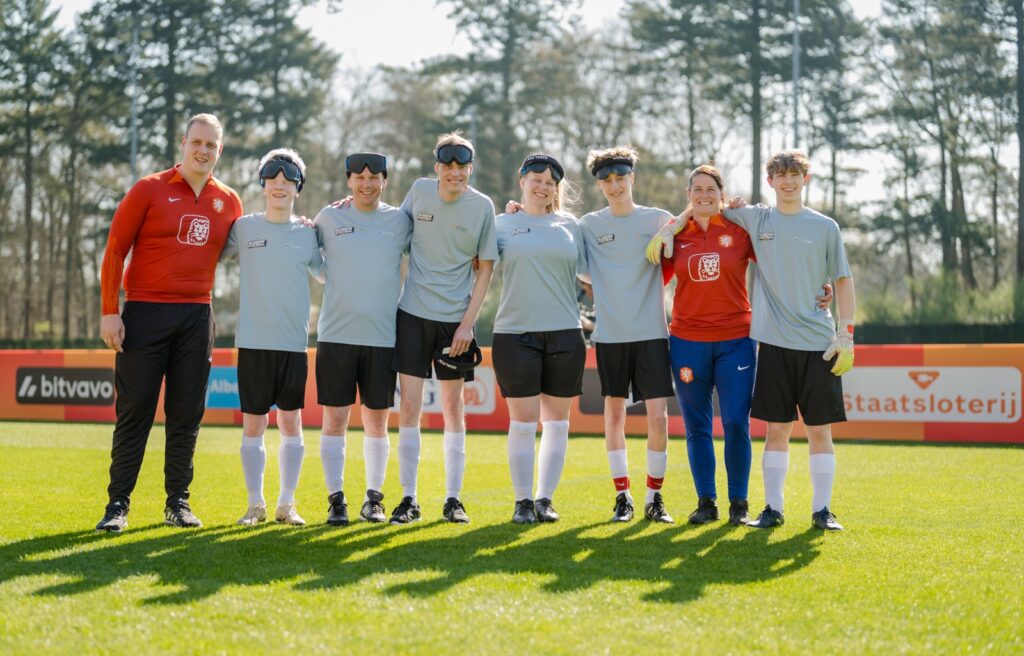 groepsfoto van blindenvoetbalteam, twee coaches en zes spelers op een voetbalveld