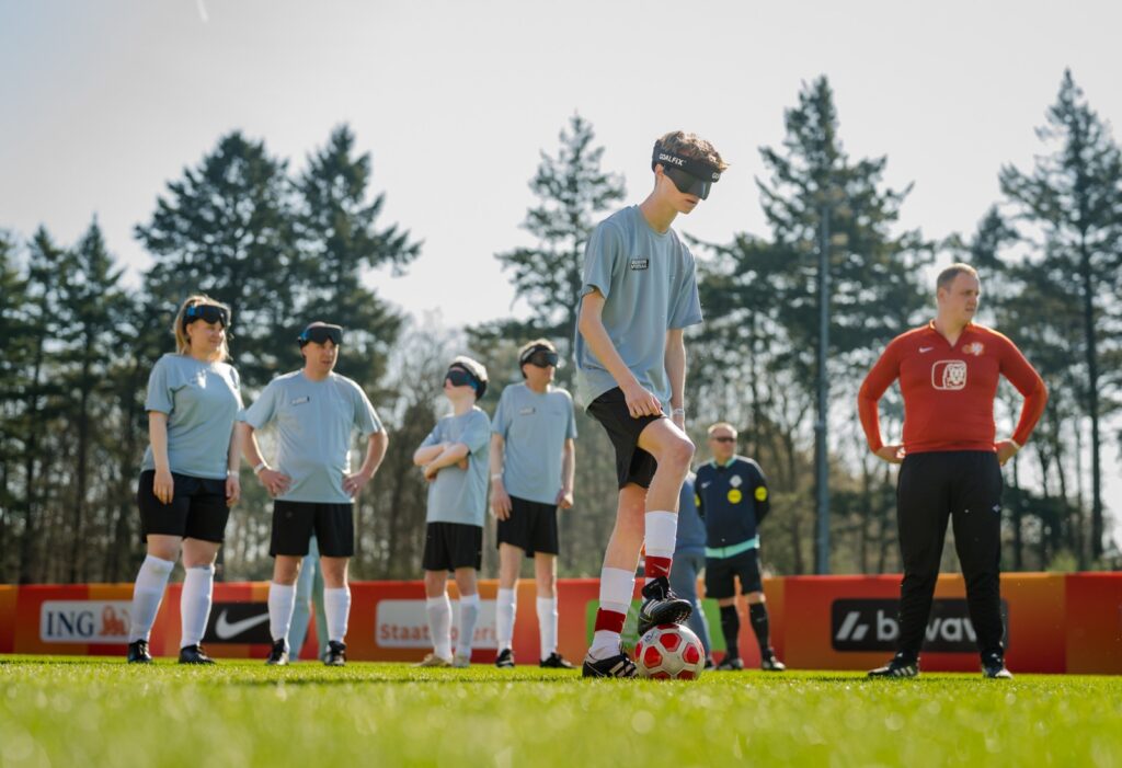 blindevoetballer wikke aan de bal tijdens een training. op de achtergrond staan teamgenoten en een trainer