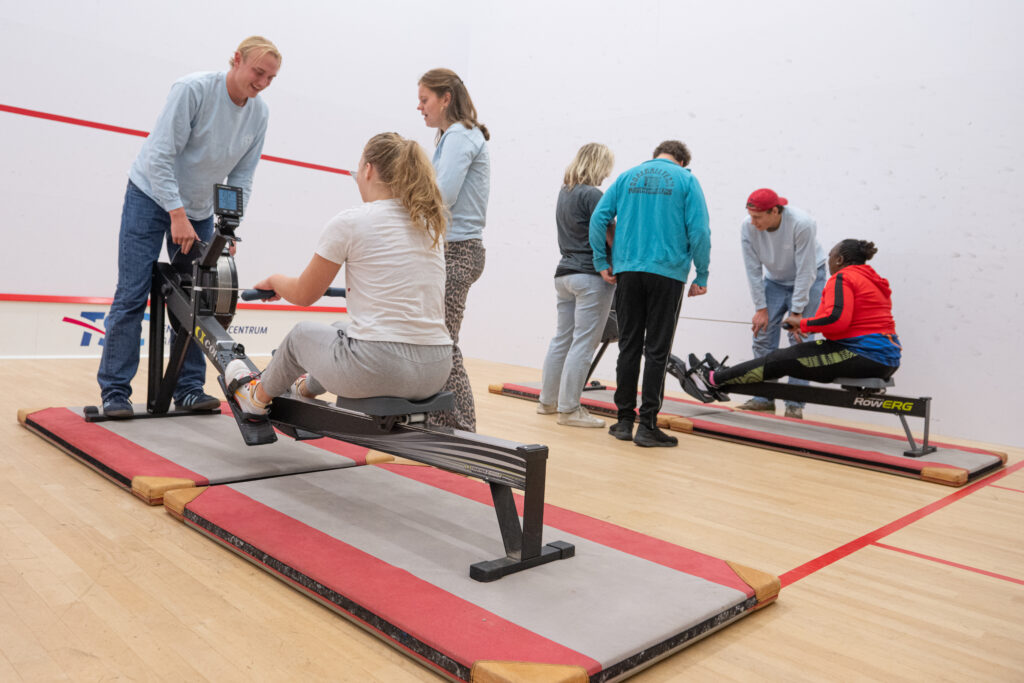 deelnemers van de multisportdagen roeien op een roeiapparaat onder begeleiding van rowing blind trainers