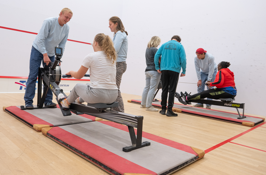 deelnemers van de multisportdag roeien onder begeleiding van rowing blind trainers op een indoor roeiapparaat